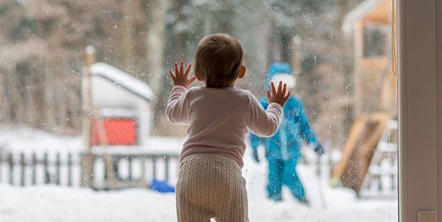 Child looking outside on a snowy day with older sibling playing outside