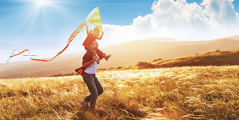 Man with kid on shoulders. Kid is holding a kite and they are running through a field