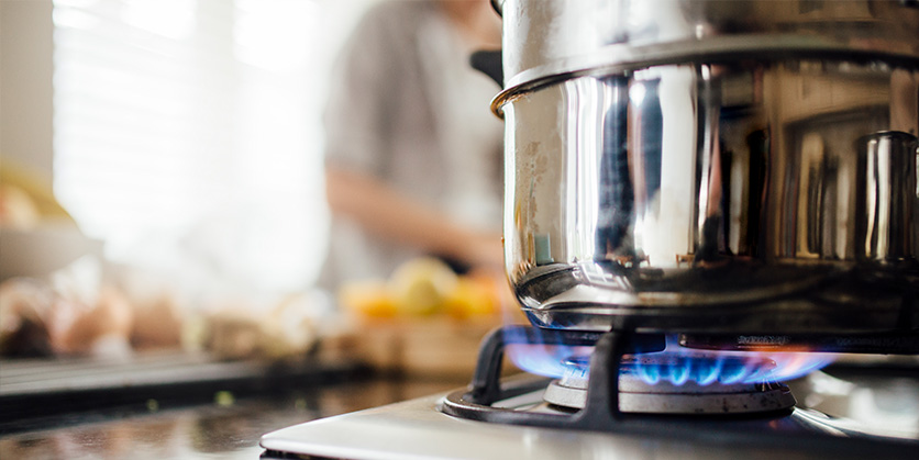 Cooking pot sitting on a gas stove, blurry person in the background chopping vegetables
