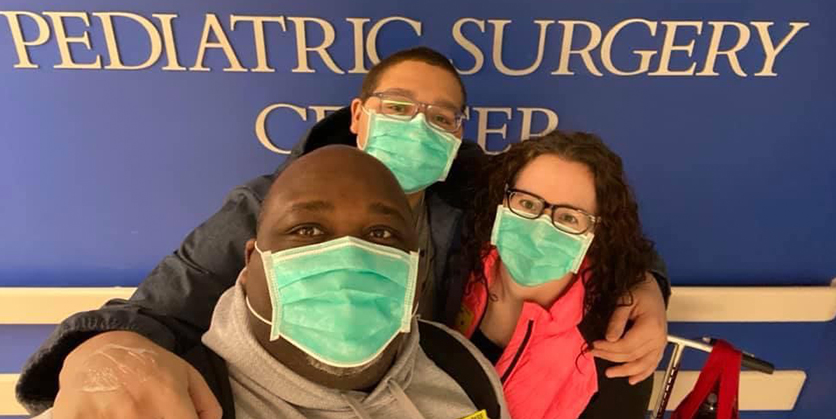 Family standing in front of pediatric surgery sign on wall, wearing masks