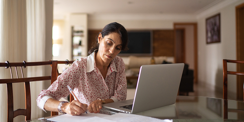 Woman sitting at table, in front of laptop, writing on some papers