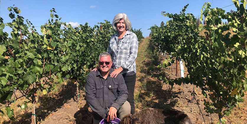 A couple poses outside, standing in their vineyard