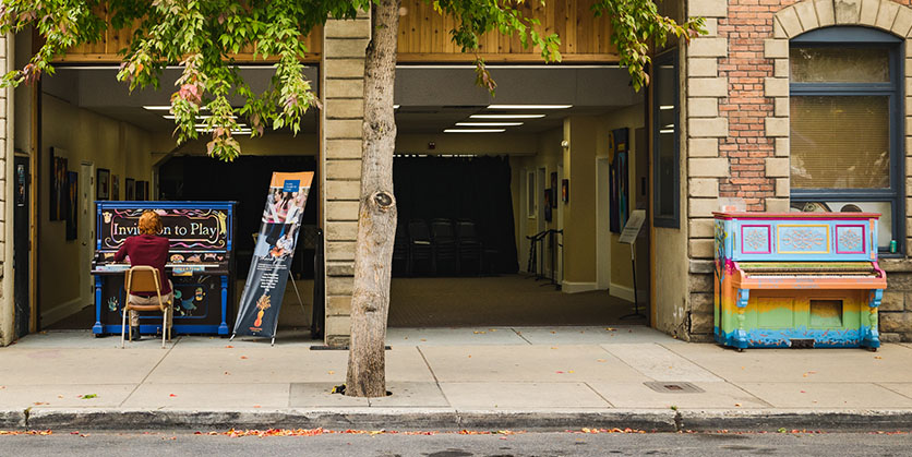 View of someone's back as they play a piano on the sidewalk. A second colorful piano is off the right next to a building