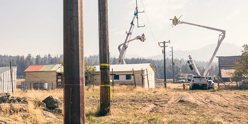 A steel transmission pole next to a wood transmission pole with workers replacing a pole in the background