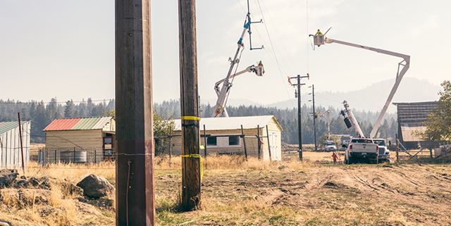 A steel transmission pole next to a wood transmission pole with workers replacing a pole in the background