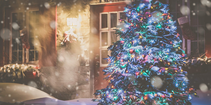 Illuminated Christmas Tree in front of a decorated home at night with snow falling