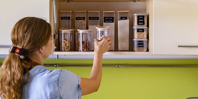 Woman placing glass jar filled with nuts in a kitchen cupboard. The cupboard is very organized with different labeled jars.