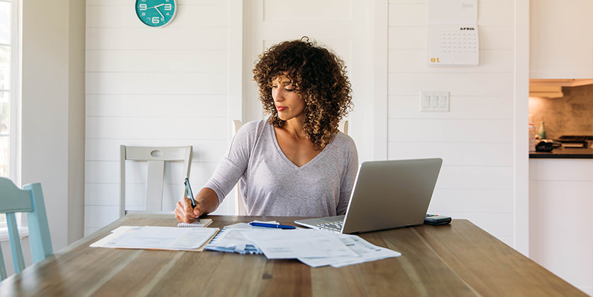 A woman sits at her dining room table with laptop and financial reports doing her monthly budget