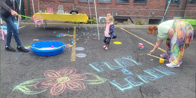 Woman drawing with chalk outside, little girl stands nearby with a mini pool and a man blowing bubbles