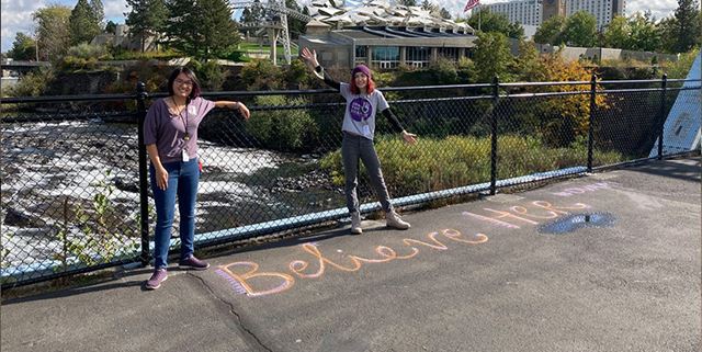 Two woman smile near a chalk drawing that reads "Believe her"