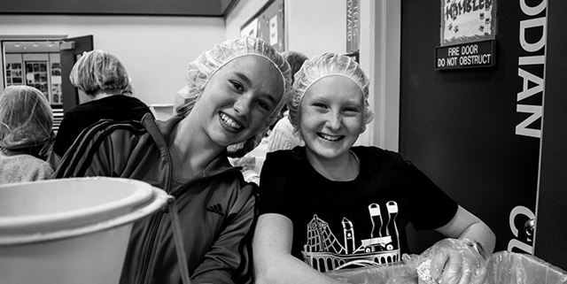 Two children wearing hair nets and smiling as they pack food