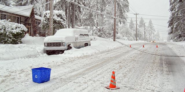 Power lines are on top of a snowed in car and on the snow covered street with traffic cones placed around the area