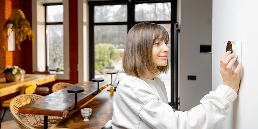Woman adjusting home thermostat, she is wearing a sweater and there is snow visible through the window