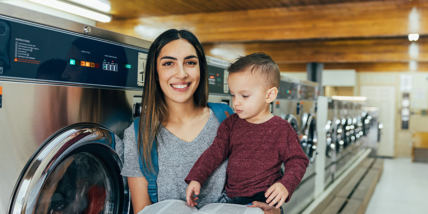 Woman wearing a backpack and holding a school book and baby stand in a laundrymat