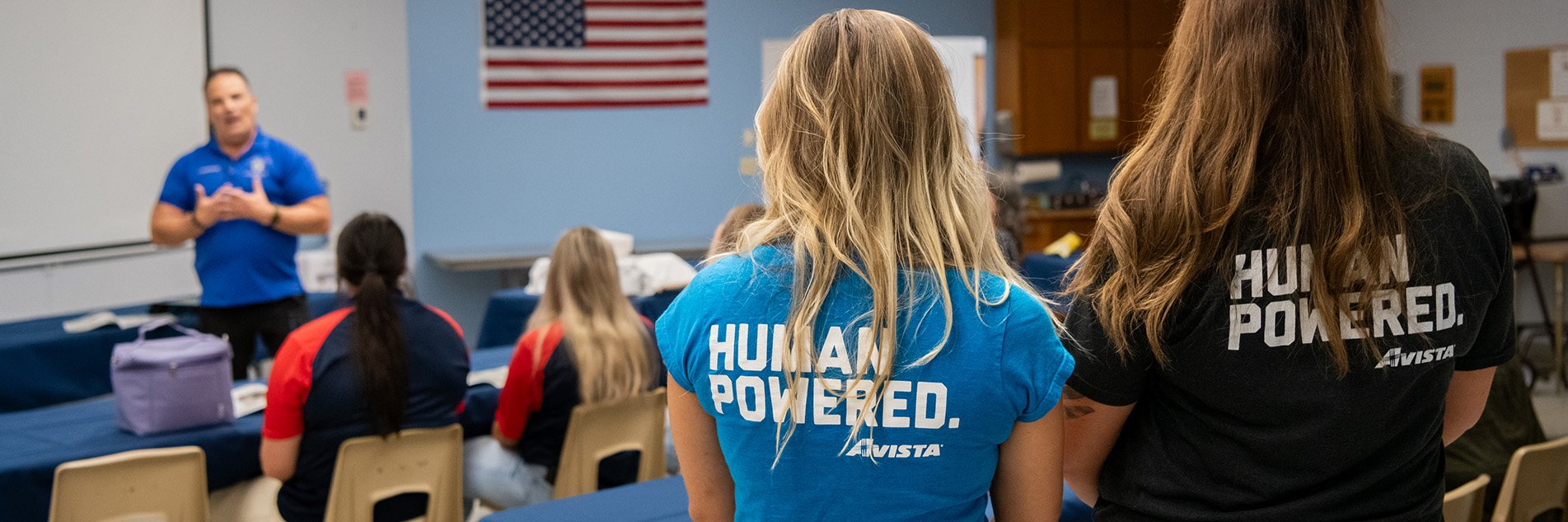 Two women stand and watch a man talking to a classroom. The women are wearing blue t-shirts that say "Human Powered. Avista."