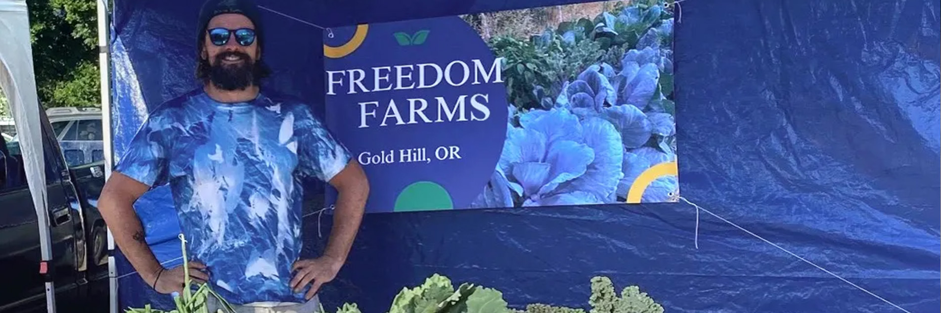 Bearded man wearing sunglasses and a blue t-shirt, smiles in front of a Freedom Farms Gold Hill, Oregon sign at a farmers market vegetable table.