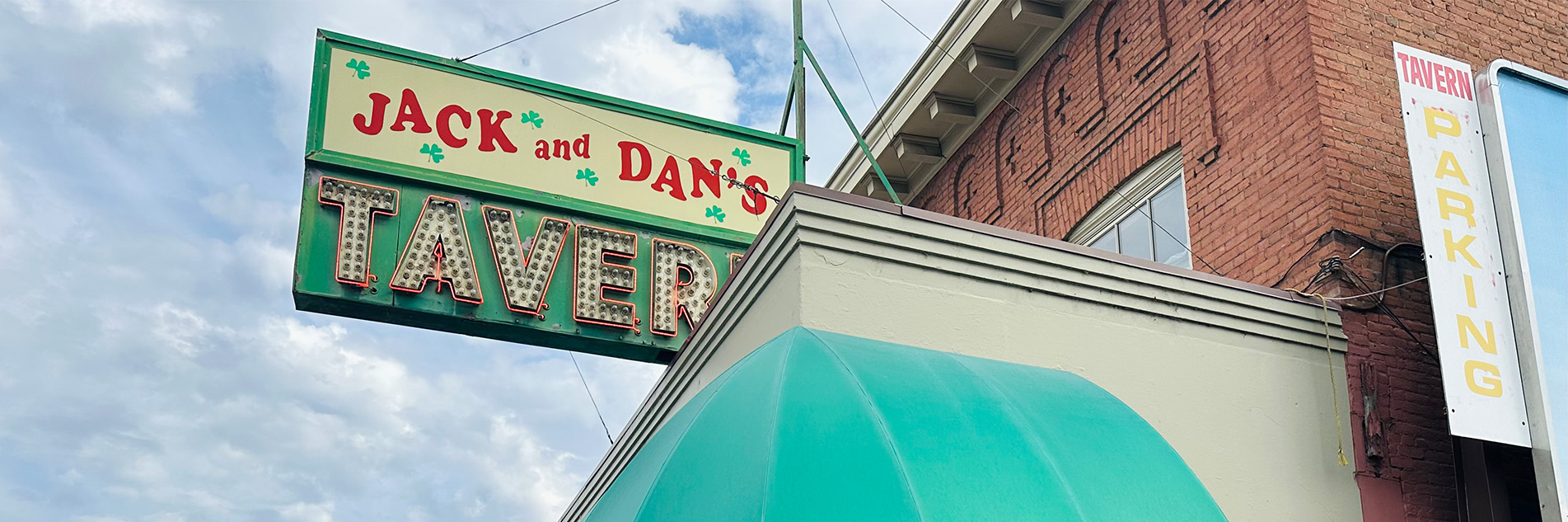 Sign on a brick building, the top half of the sign says Jack and Dan's in bold red writing on a white background with green three-leaf clovers, the bottom half of the sign says tavern in white lettering with lights  and a red lighted border on a green background.