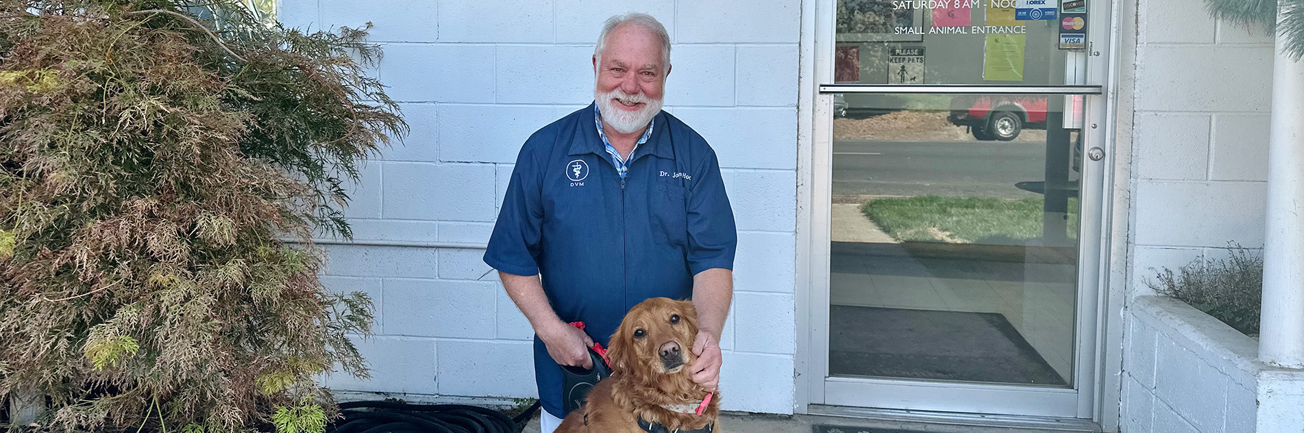 Man with white hair and a white beard and moustache, wearing a navy blue veterinary jacket, kneels down next to a golden retriever dog in front of a white brick building.