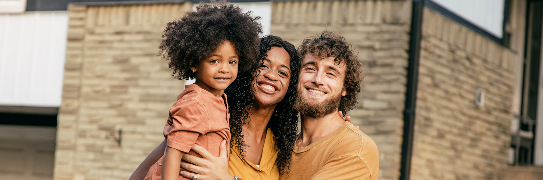 Mother and father holding child outside in front of a building, they are all smiling and look happy.