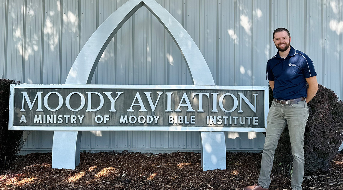 Smiling man standing outdoors next to a sign that says Moody Aviation a ministry of Moody Bible Institute.