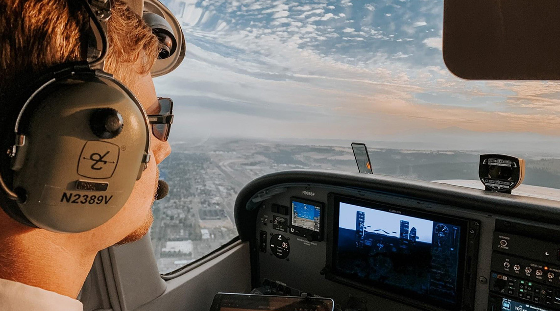 Looking out the window of the cockpit of a propeller airplane as a man in sunglasses flies it over the city during a cloudy day.
