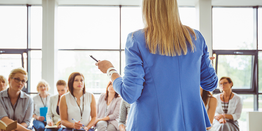 Blonde-haired woman wearing a blue blazer is standing giving a presentation in front of a group of sitting professionals.