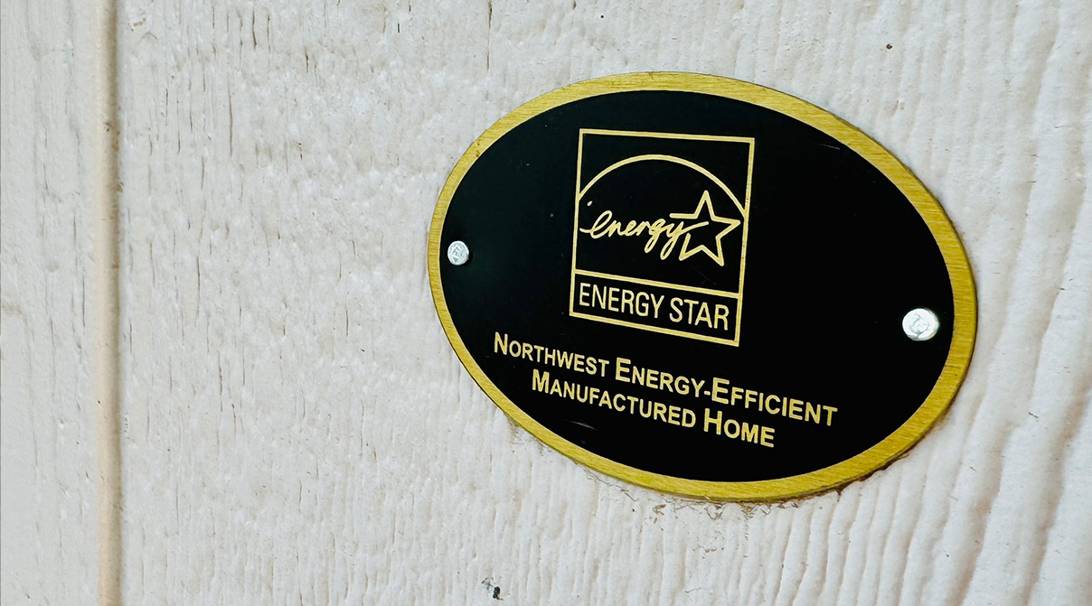 Closeup of a circular sign attached to wood siding, the sign is black and gold with the Energy Star logo and the words Northwest Energy-Efficient Manufactured Home.