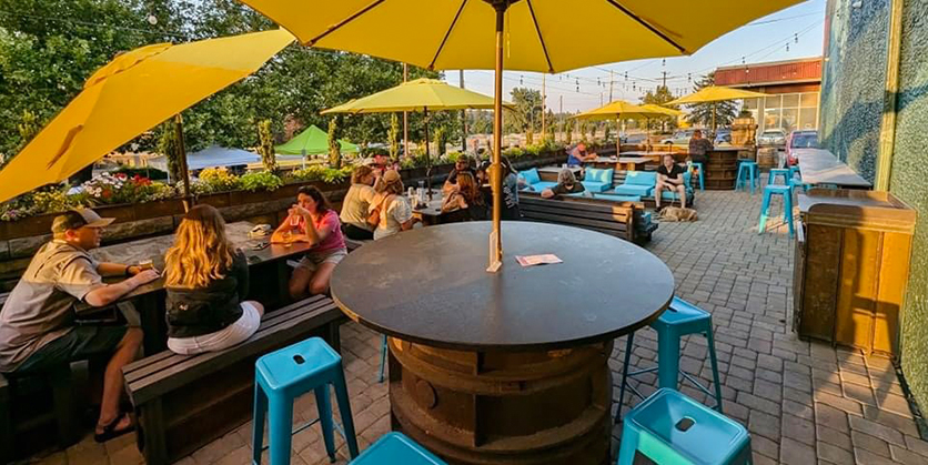 Outdoor patio with groups of people enjoying a drink at tables that have yellow umbrellas and blue stools.
