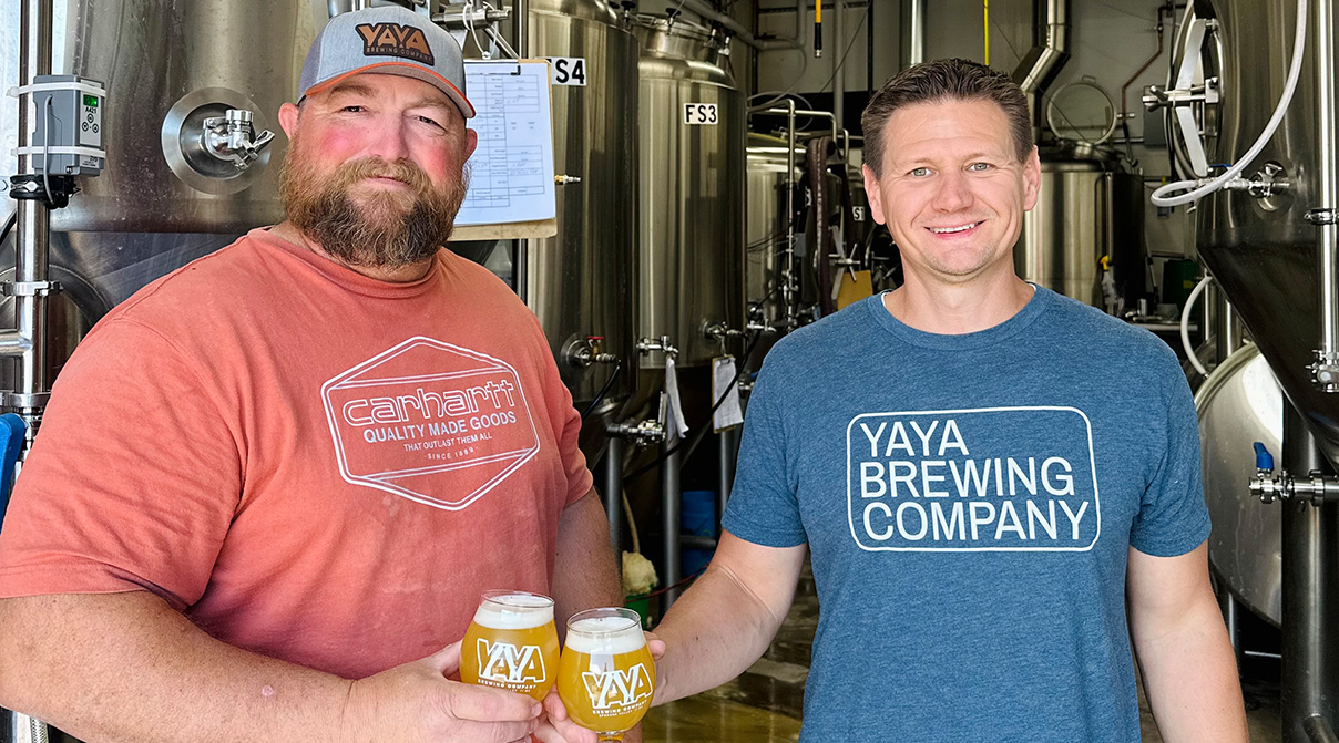 Two men stand inside a commercial brewing room, they are both holding glasses of freshly poured beer from YAYA Brewing Company.