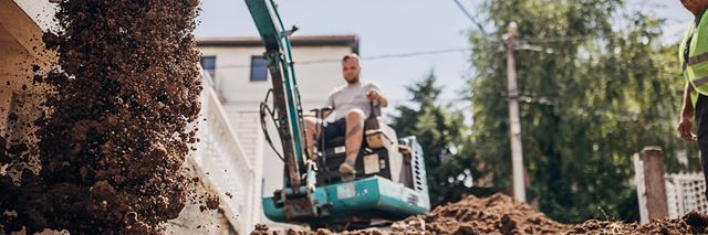 Man using a teal excavator to move dirt outside.