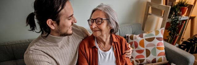 Man has his arm wrapped around the shoulder's of his grandmother while they sit on the couch, smiling at each other.