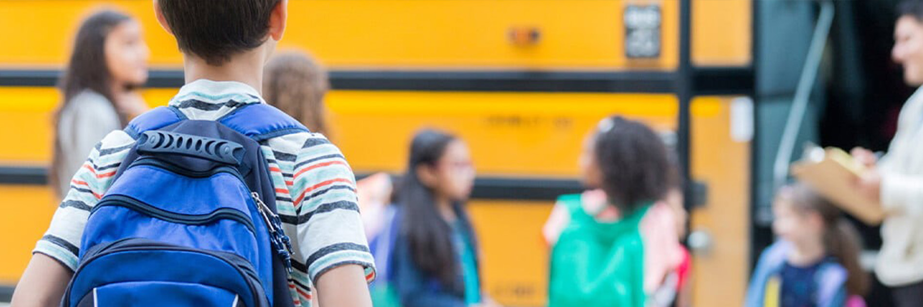 Boy wearing a blue backpack as he lines up to get on the school bus, other kids with backpacks are in front of him while the teacher checks off attendance by the bus doors.