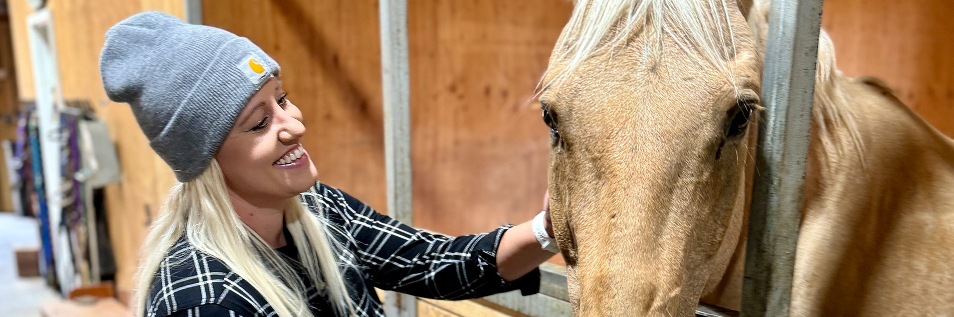 Smiling woman pets a horse while it stands in a stable stall.