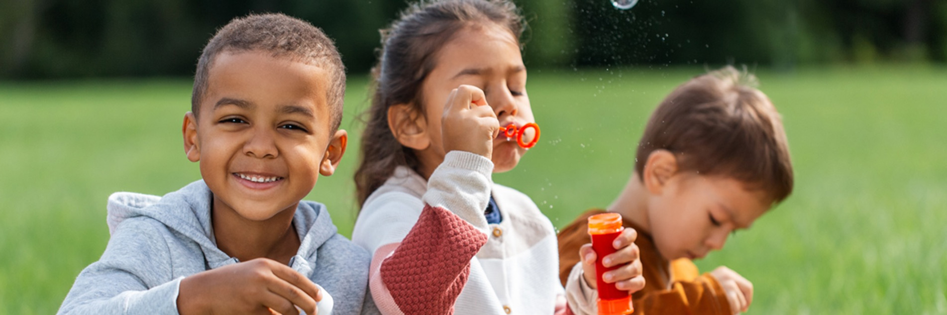 Three children sitting on the grass outside blowing bubbles.