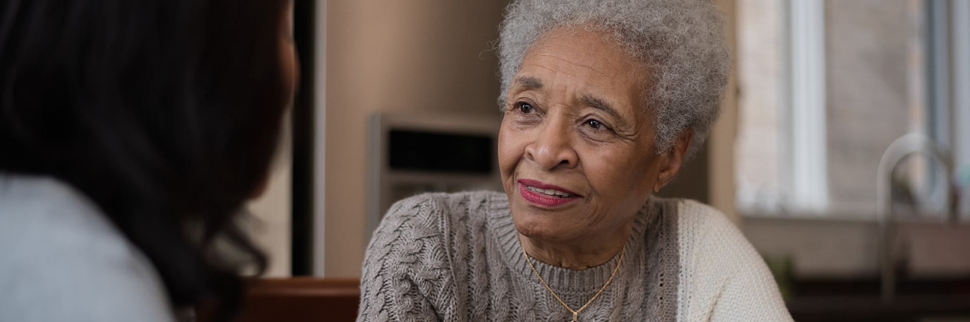 Senior woman sitting with another woman at a kitchen table, chatting and drinking coffee.