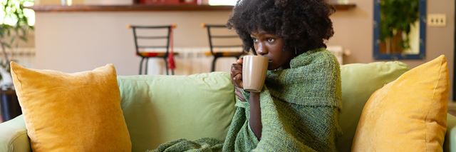 Woman sitting on couch, wrapped up in a blanket, drinking a warm drink out of a mug.