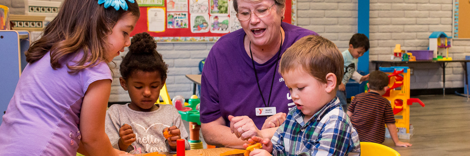 YMCA staff member smiles, crouched down next to a table of three preschool children playing with Play-Doh.