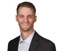 Smiling man with brown, short hair with a bit of facial hair growing in on his face, wearing a black suit jacket and a white button up shirt.