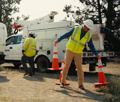 Person in yellow Avista vest and white hardhat places safety cones behind two Avista trucks outdoors.