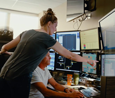 Man sits at desk at work with 8 screens, a woman is standing next to him and is pointing to one of the screens.