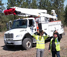 Two men in yellow safety vests stand outside in front of an Avista truck, one is looking through binoculars and one is pointing upwards.