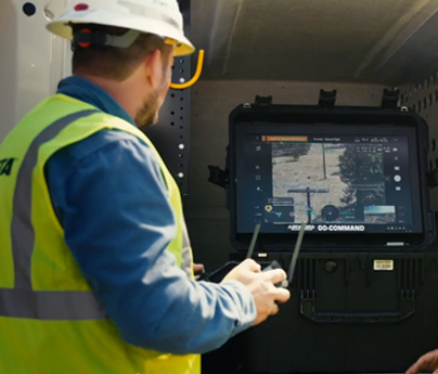 A man wearing a yellow safety vest looks at a screen while using a drone controller to pilot a drone.