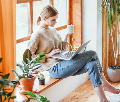 Woman on laptop, holding a mug in one hand, sitting in a sunny room with plants