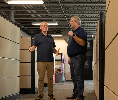 Two men are standing and chatting between cubicles in the office.