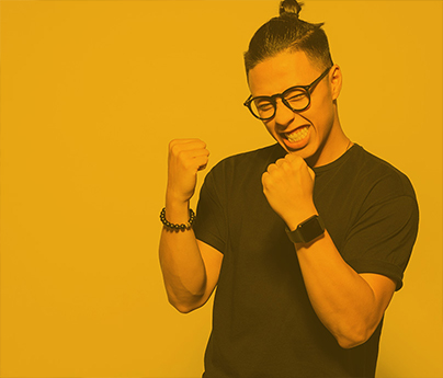 Man with dark hair pulled up in a bun, wearing black glasses and a black t-shirt stands against a yellow background. The person has a huge smile with his arms bent at the elbows, with fists near his chin, in a pose of excitement.