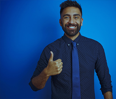 Man with dark hair and a short beard and moustache, wearing a navy blue long sleeve shirt with white dots and a navy blue tie stands in front of a solid blue background. He is smiling and making a thumbs up sign with his hand.