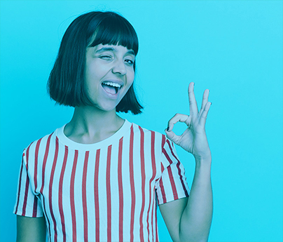A woman is winking, with her hand held up, her pointer finger and thumb touching to make a circle while her other fingers are pointing into the air in an "OK" sign as she stands in front of a blue background.