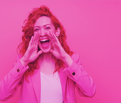 Smiling woman has her hands up to her mouth like she is shouting excitedly, standing  in front of a pink background.