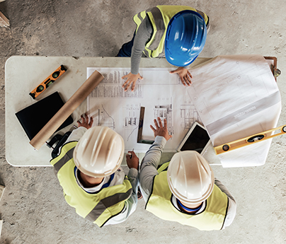 People in safety vests and hard hats look at a blueprint spread out on the table, they are pointing to it as they discuss it.
