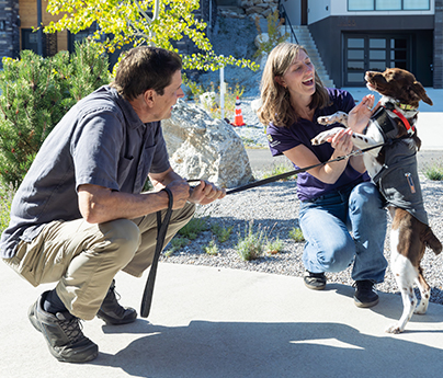 Man walking a dog is crouched down, watching the dog say hello to a woman who is crouched down, smiling and petting the dog.